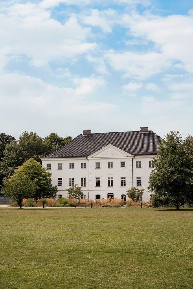 A grand white manor house with symmetrical windows sits on a manicured lawn under a bright blue sky with puffy clouds.
