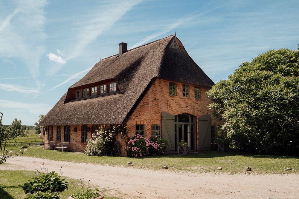 Traditional brick farmhouse with thatched roof and green landscaping stands under blue summer sky on rural property.