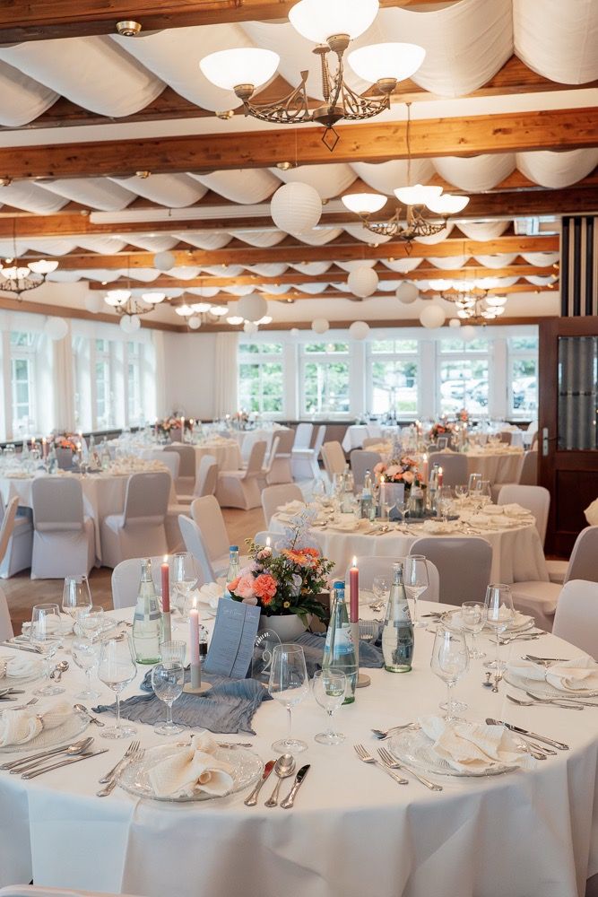 Elegant wedding reception venue with round tables, white linens, chandeliers and exposed wooden beams in rustic ballroom.