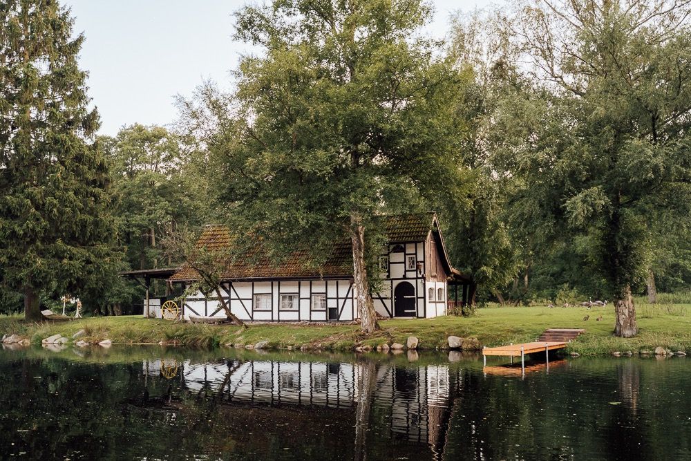 Scenic panoramic view of a tranquil lake surrounded by autumn trees and rustic buildings in a peaceful countryside setting.