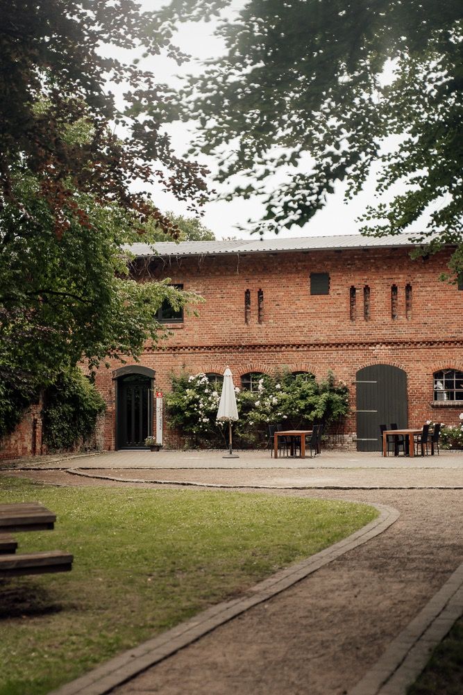 A panoramic view of a rustic brick building with outdoor seating and dining tables in a courtyard surrounded by trees.