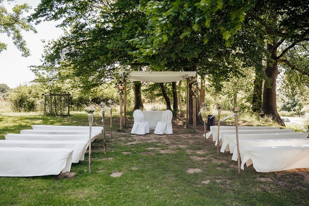 Outdoor wedding ceremony setup with white chairs and wooden pergola under shady trees in a garden setting.