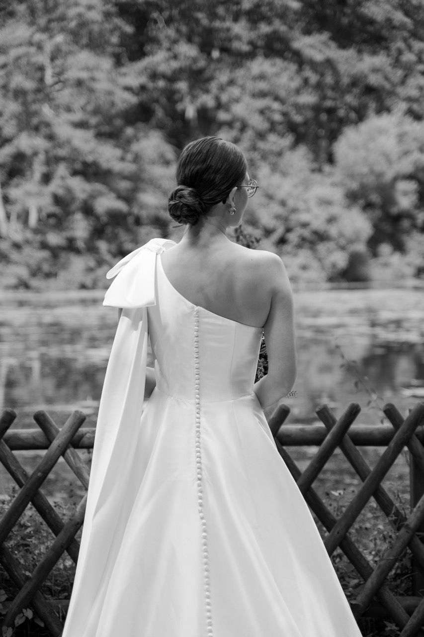 Black and white photo of a bride in an elegant one-shoulder wedding gown with bow detail, standing by a wooden fence.