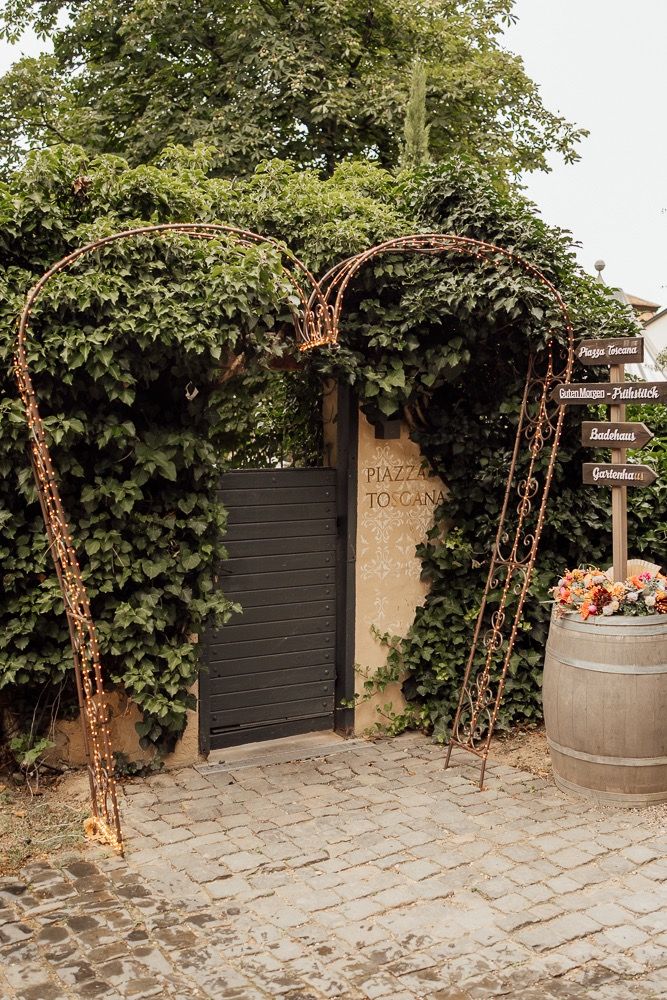 Rustic outdoor venue with wooden archway, wine barrel, and climbing vines against a stone wall and black gate.