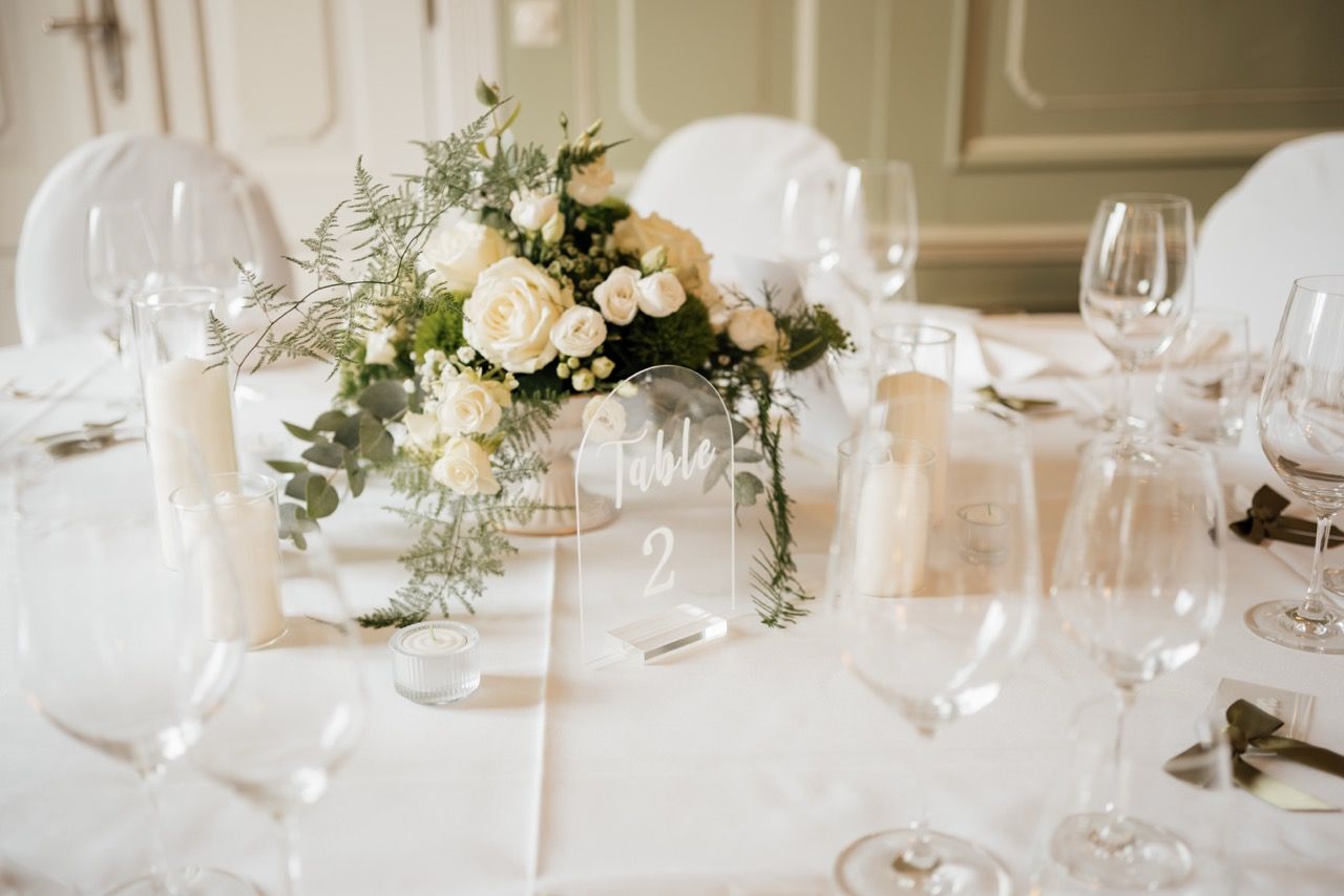 Elegant wedding table setting with white roses, greenery centerpiece, candles, and crystal glassware on white linen.