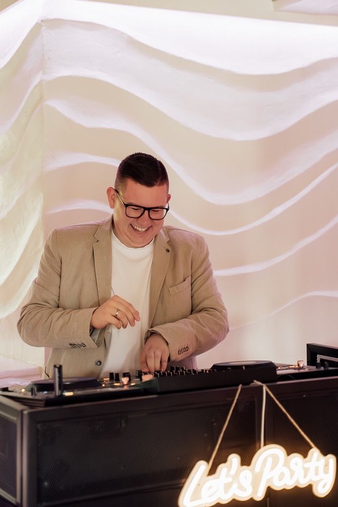 DJ in beige blazer and glasses performs at event with white wavy backdrop and illuminated signage.