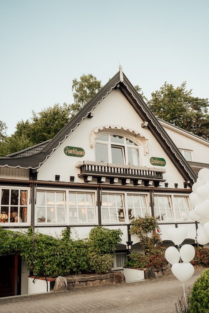 White rustic wedding venue building with traditional architecture and large windows set against green trees and foliage.