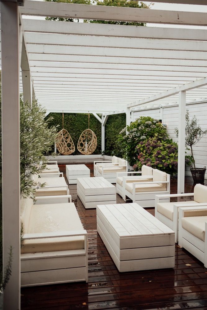 Modern white outdoor lounge area with multiple daybeds arranged on wooden decking under a pergola structure.