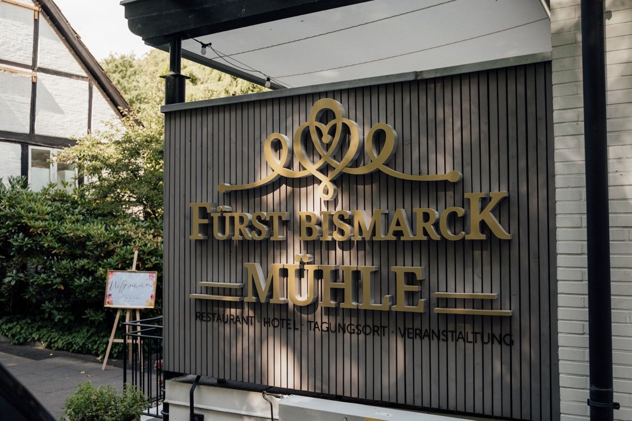 Gold lettered Furst Bismarck Muhle sign mounted on a dark corrugated metal wall surrounded by lush greenery.