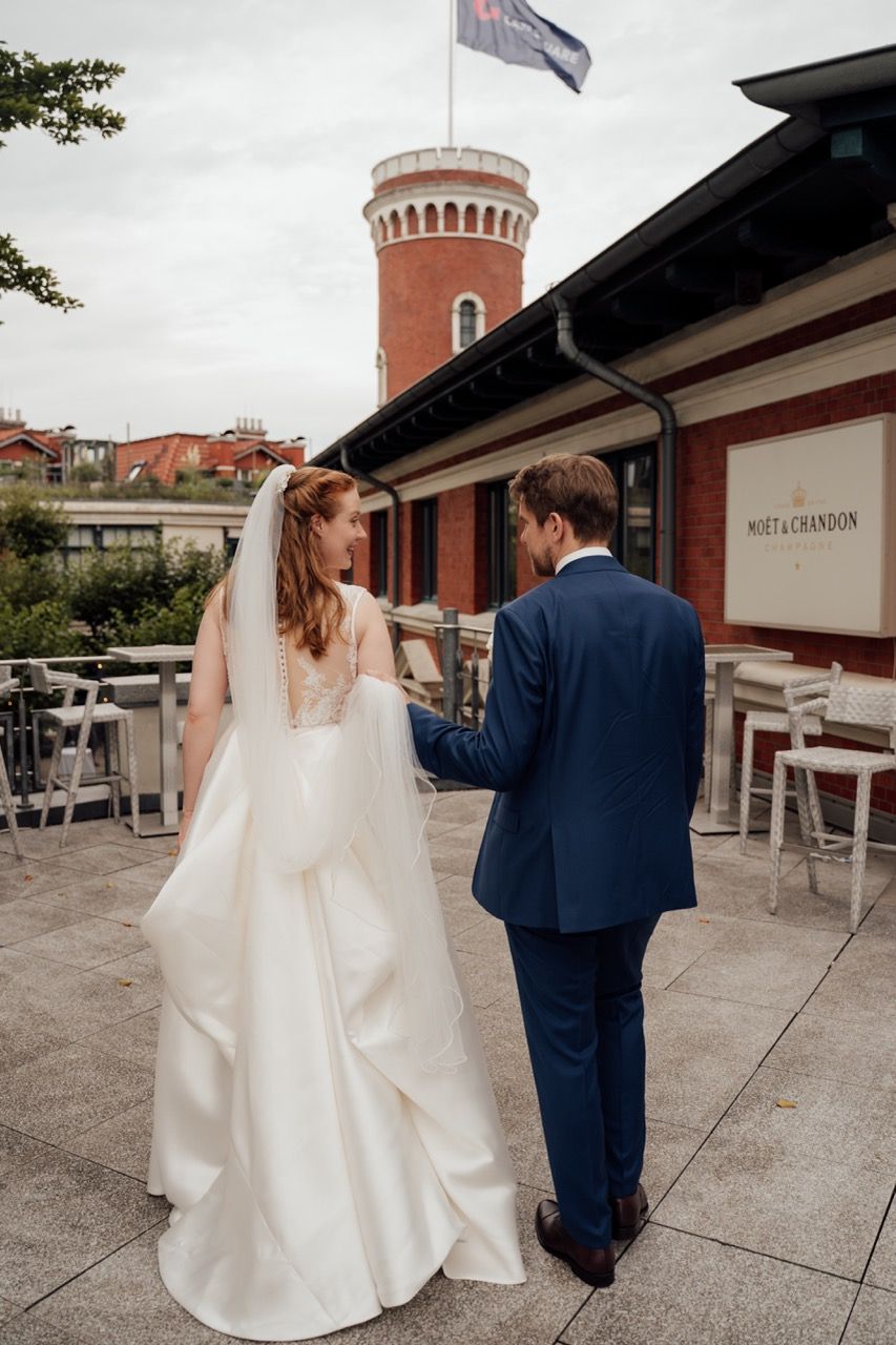 Bride and groom walking hand in hand on a terrace near a red brick tower on their wedding day.
