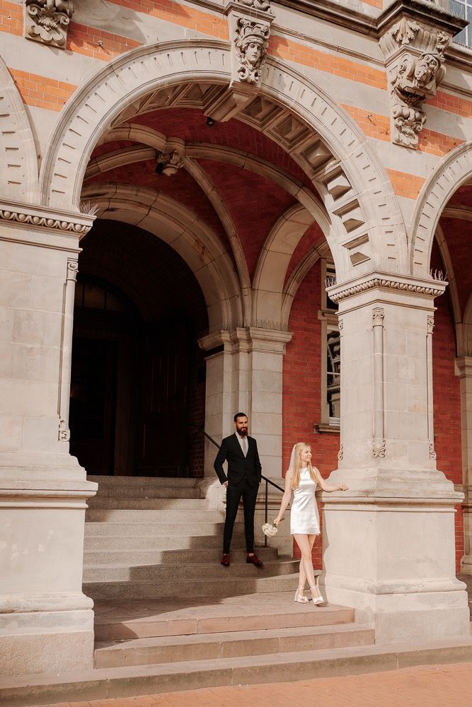 A couple stands beneath ornate stone archways of a historic building in a romantic architectural setting.