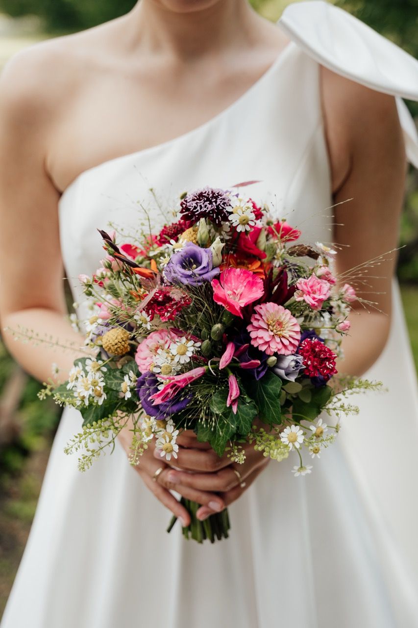 Bride in white one-shoulder gown holding a vibrant wildflower bouquet with pink, purple, and red blooms.