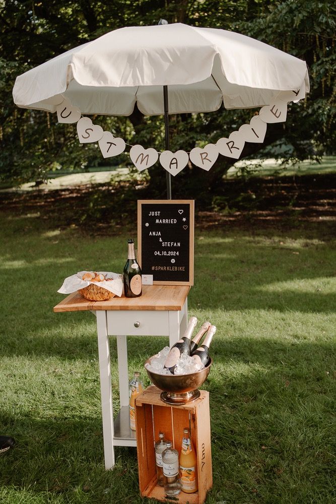 Rustic wooden beverage station with white umbrella and chalkboard sign set up on green lawn for outdoor event service.