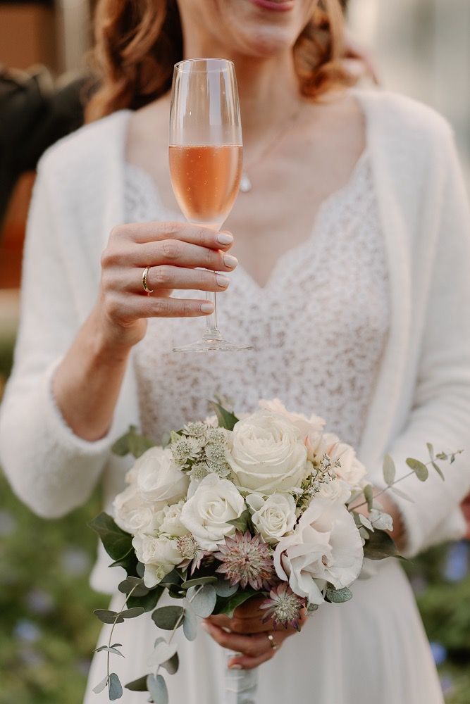A bride in white lace holds a glass of rose champagne and a delicate bouquet of white roses and eucalyptus.