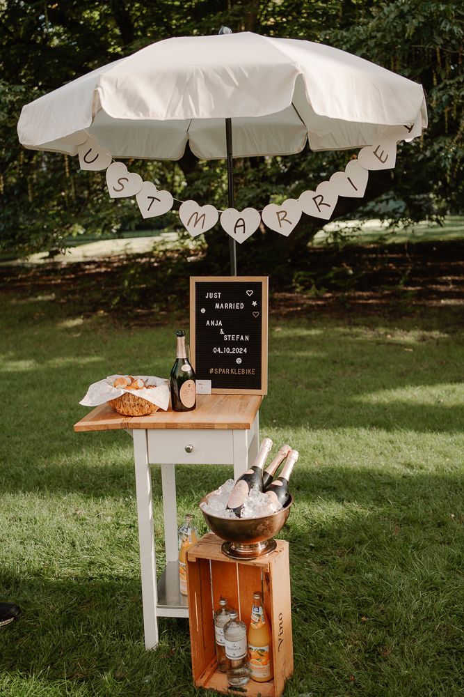 Rustic outdoor beverage station with white umbrella and wooden crate displaying drink dispensers on a sunny lawn.