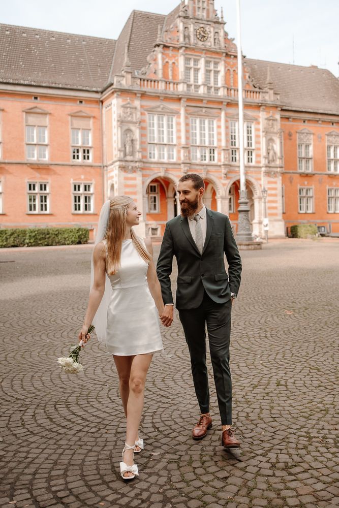 A couple walks hand in hand on cobblestone pavement in front of a grand historic European-style building.