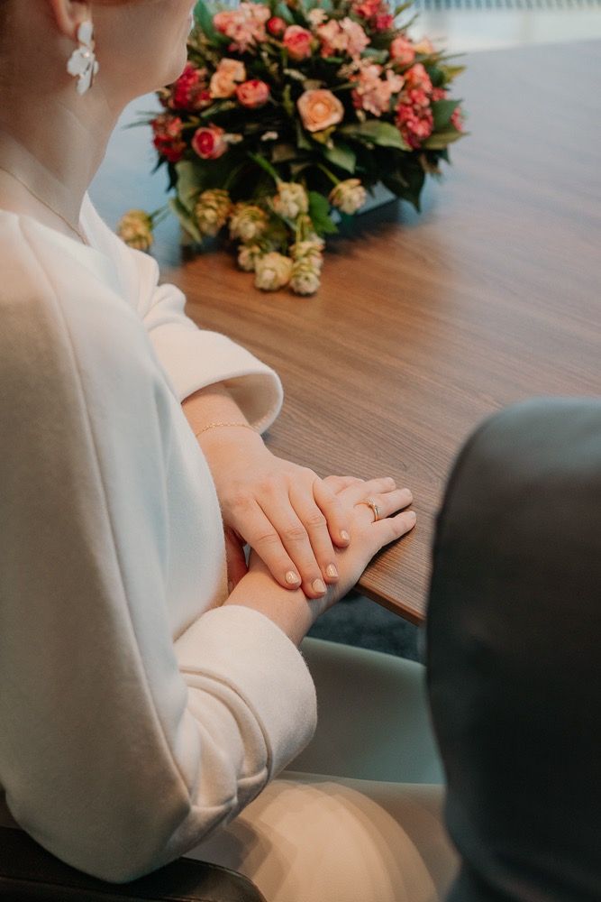 Close-up of clasped hands wearing a wedding ring with a colorful floral bouquet in the background on wooden surface.