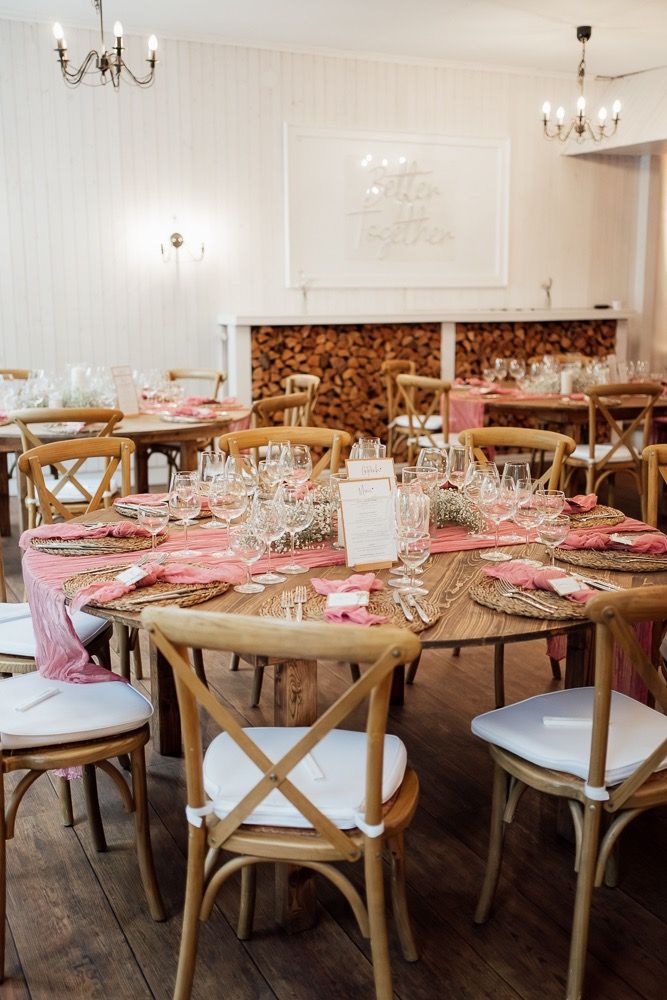 Elegant wedding reception tables set with rustic wooden chairs and pink floral centerpieces in a bright white room.