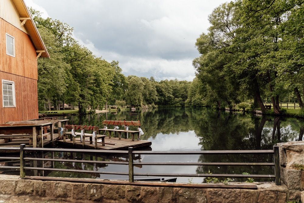 A panoramic view of a wooden dock extending into a tranquil lake surrounded by lush green trees on a cloudy day.