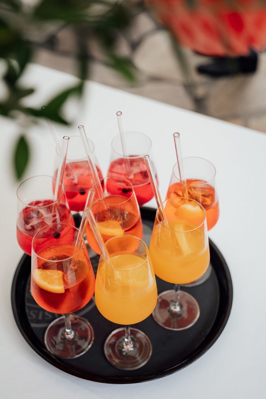 A tray of colorful cocktails with straws, featuring red and orange drinks garnished with fruit slices and ice.