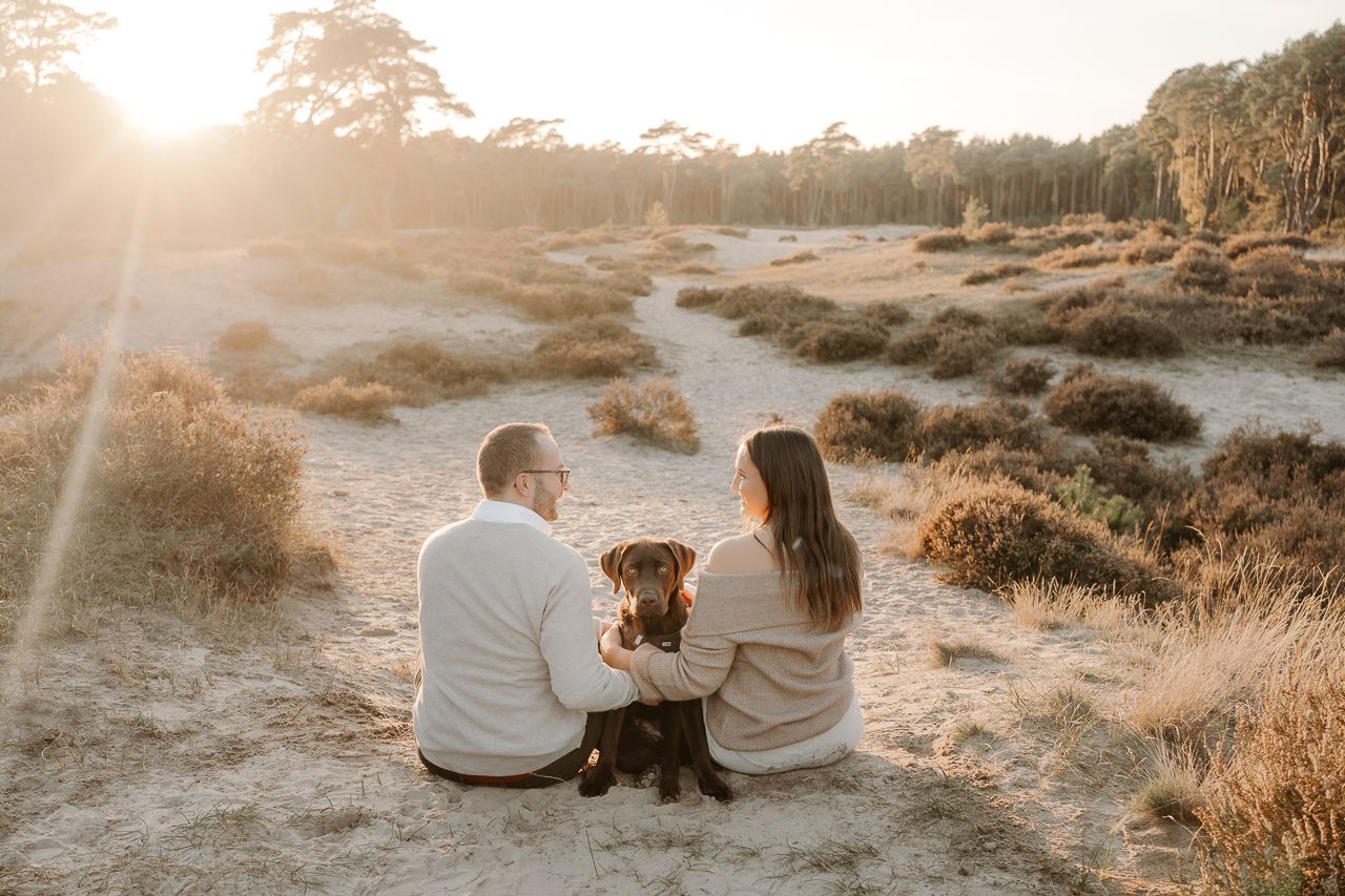 Ein Paar sitzt mit seinem Hund auf sandigem Boden während der goldenen Stunde bei Sonnenuntergang.