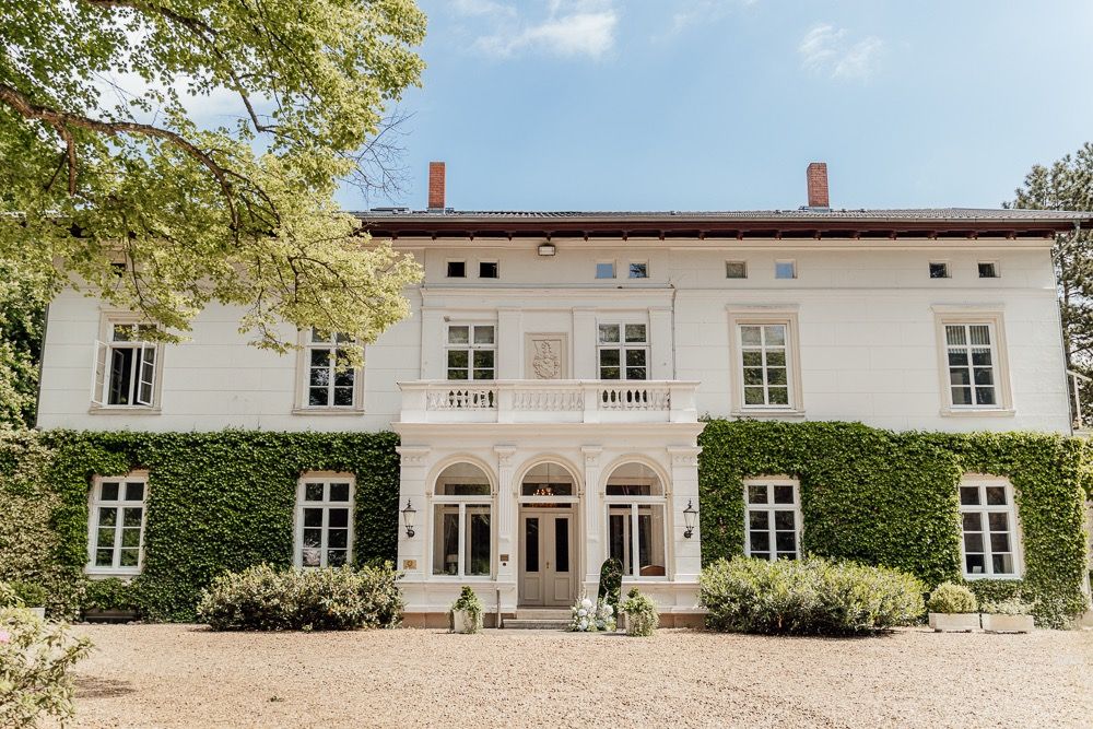 Elegant white mansion with ivy-covered walls and arched doorways sits along a gravel driveway on a sunny day.
