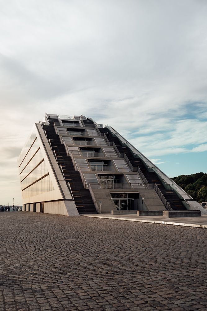 Modern angular building with dark geometric facade sits dramatically against cloudy sky on cobblestone ground.