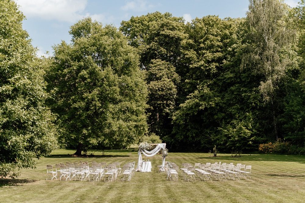 Wide view of a garden wedding venue with rows of white chairs arranged on a grassy lawn surrounded by trees.