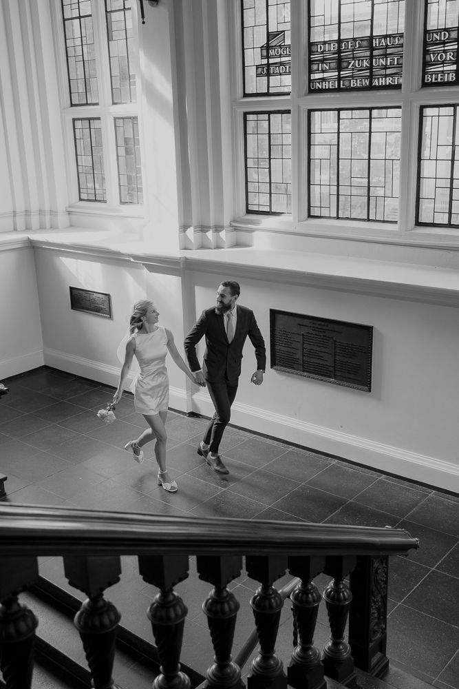 A couple in formal attire runs up a staircase together in a black and white photograph with large windows.