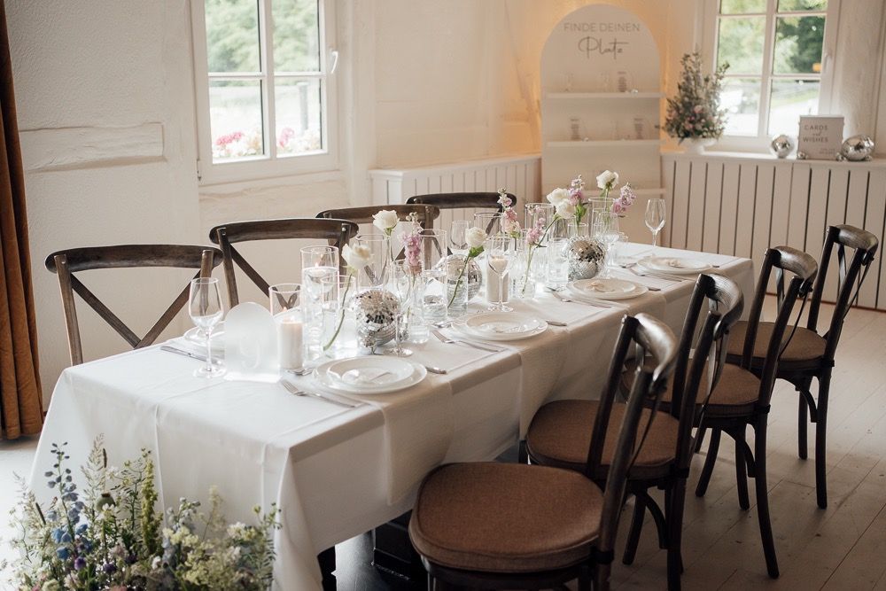 A bright reception venue with rustic wooden chairs and white tablecloths decorated with simple floral centerpieces.