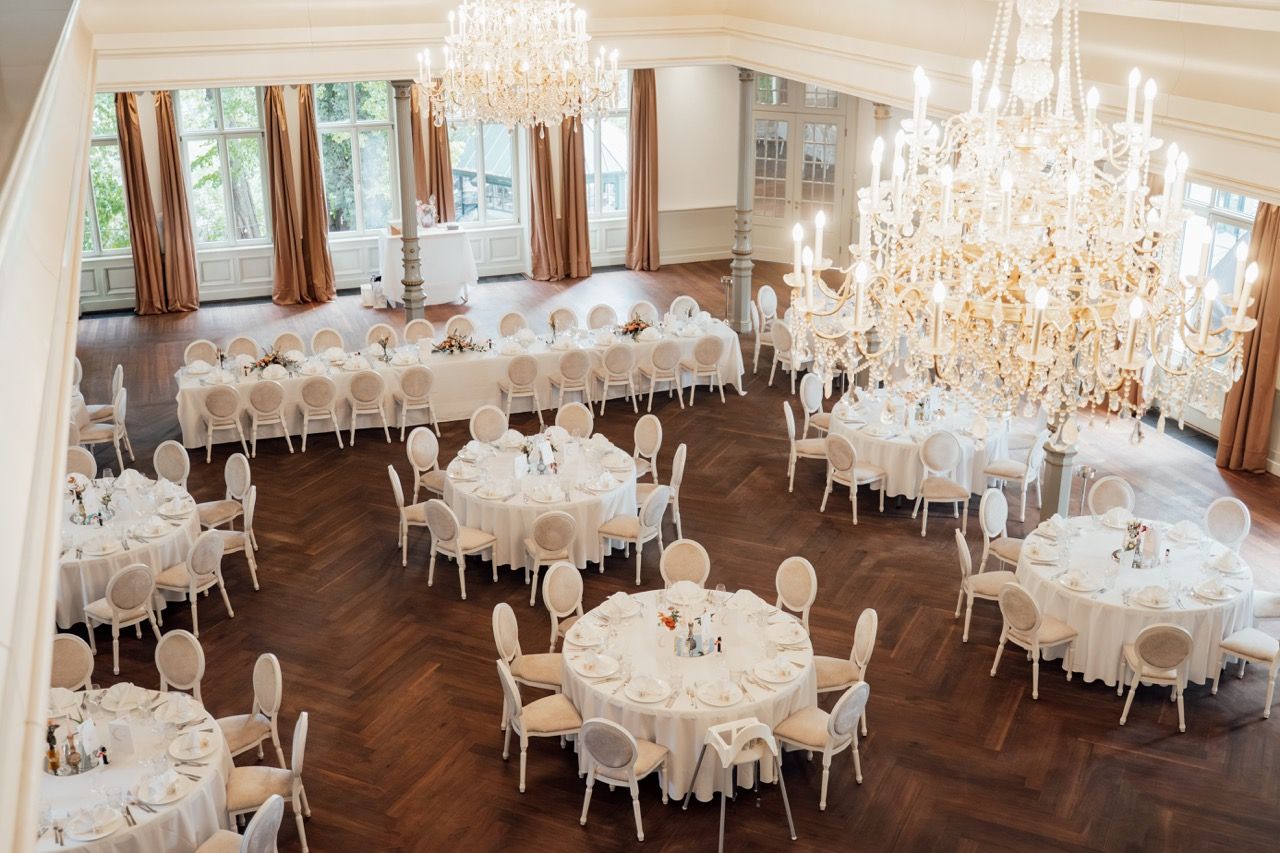 Elegant wedding reception hall with round tables, white linens, ornate chandeliers, and herringbone hardwood floors.