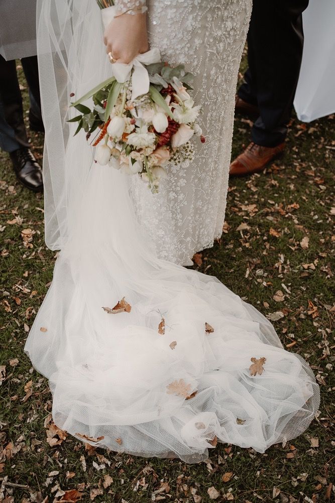 Delicate white wedding dress train flows over autumn leaves on the ground as butterflies rest on the tulle fabric.