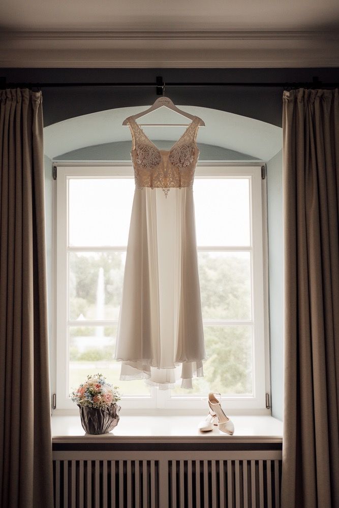 A delicate blush-colored dress hangs in a window with brown curtains and natural light streaming in.