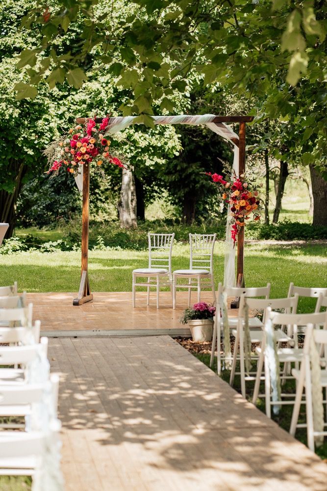 Outdoor wedding ceremony setup with white chairs and red floral arch decorations in a garden setting.