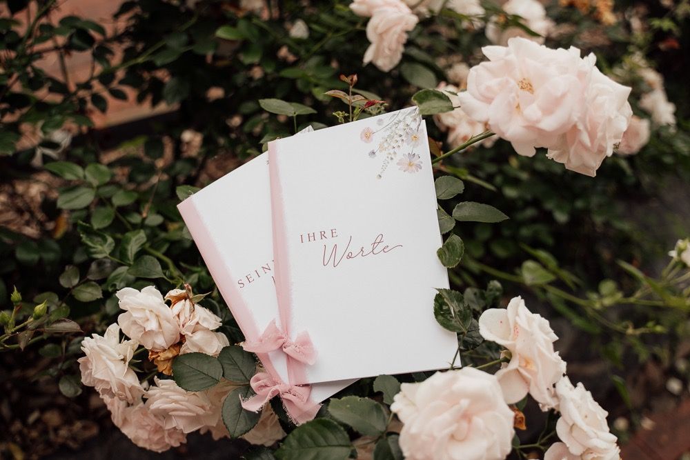 Delicate blush pink floral arrangements and wedding stationery styled on a dark textured surface with soft lighting.