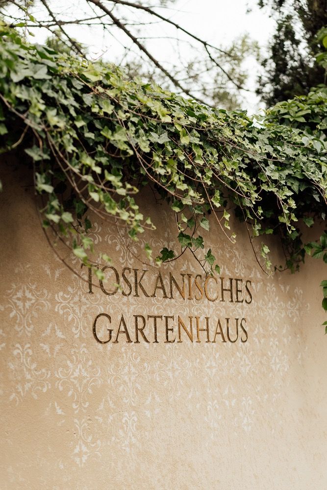 Decorative text reading 'Toskanisches Gartenhaus' on a beige wall with green ivy vines growing above.