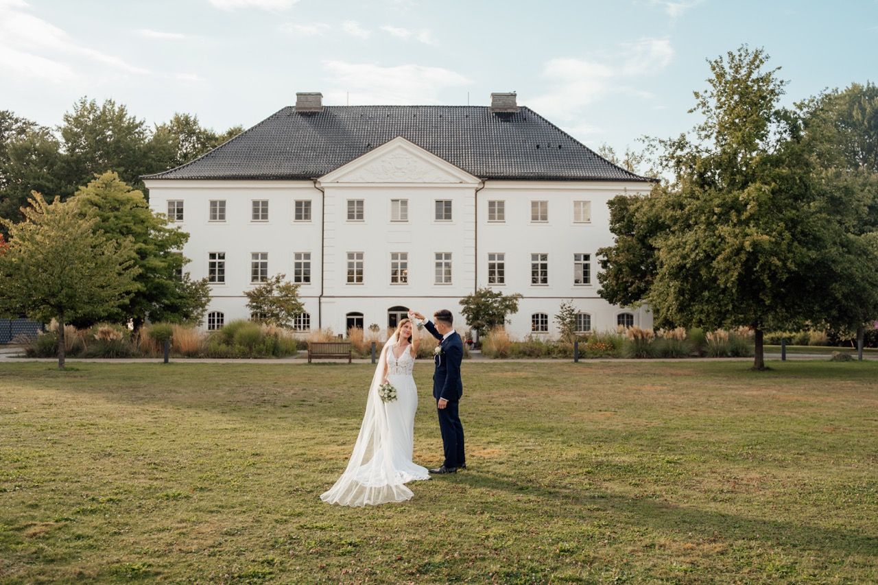 Bride and groom standing together on green lawn before elegant white manor house on sunny wedding day.
