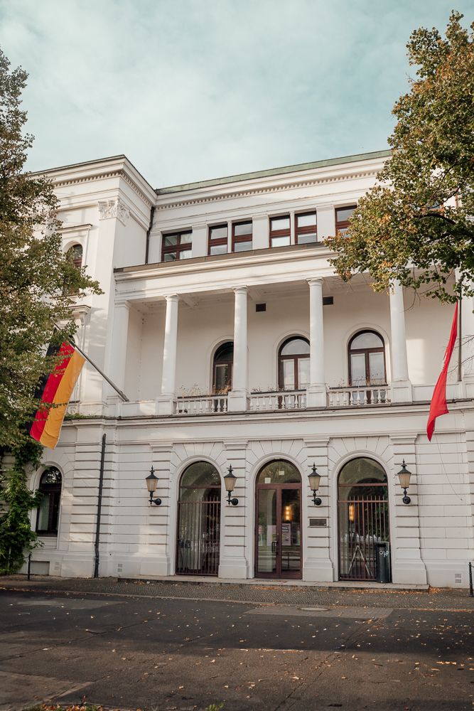 White neoclassical building with arched windows and columns displaying German and red flags at its entrance.