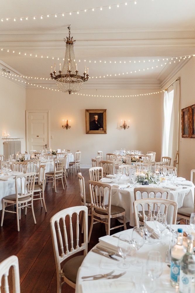 Elegant white wedding reception venue with string lights, chandeliers, and round tables set for a formal dinner.