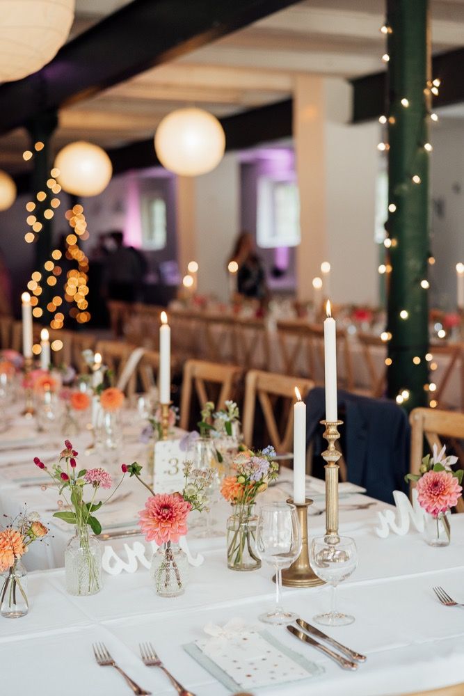 Elegant wedding reception table setting with pink dahlias in small vases and gold candlesticks on white tablecloth.