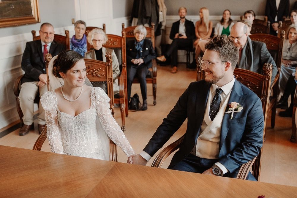 A couple signs documents at a wooden desk during their wedding ceremony while guests watch from behind.