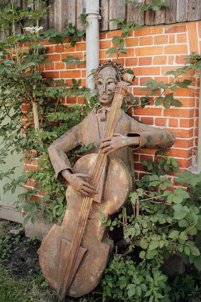 Rustic metal garden sculpture of a musician playing cello displayed against brick wall with climbing vines.