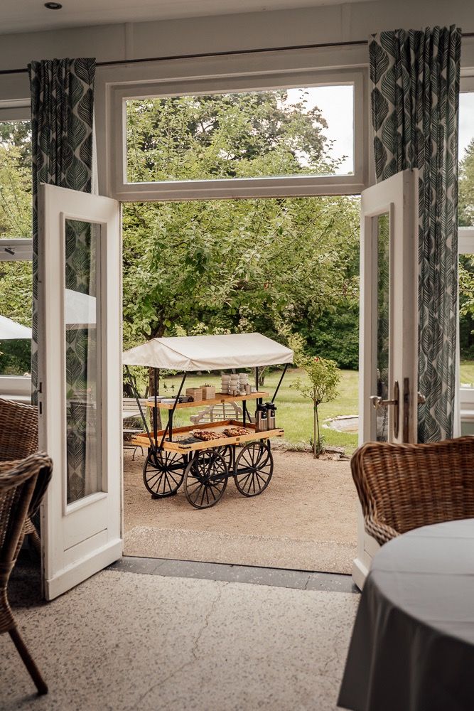 Rustic covered porch with wicker furniture and antique wagon displays overlooking a garden landscape.