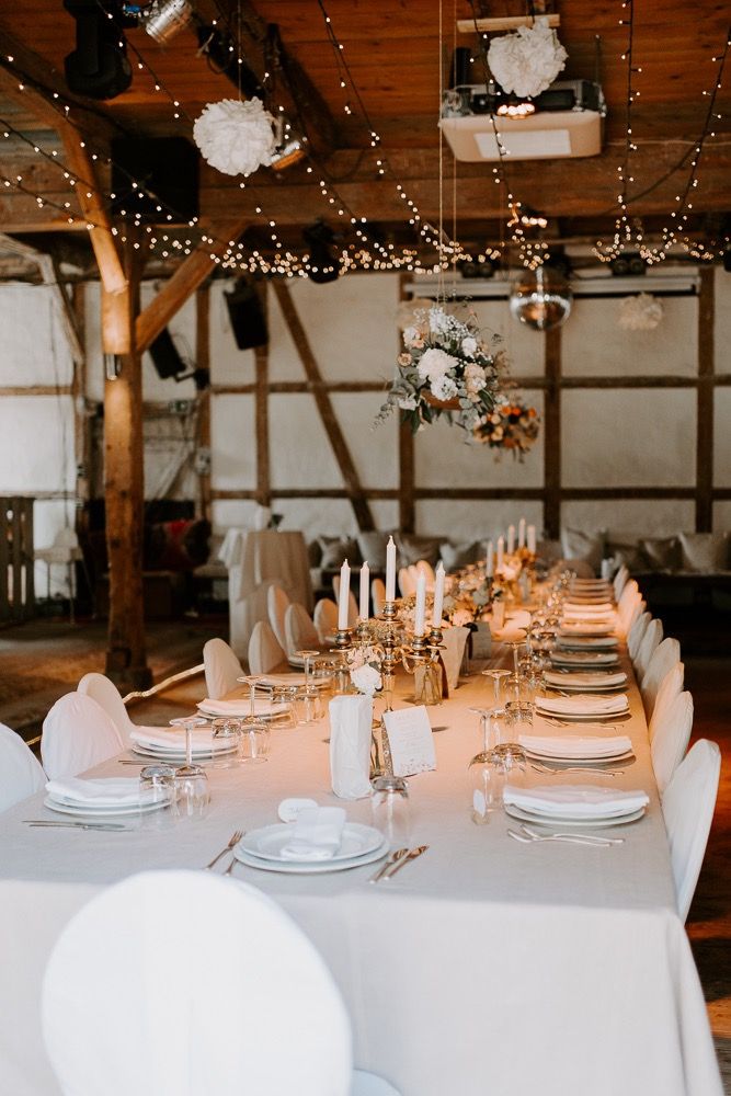Elegant table settings with white linens and candlesticks in a rustic barn venue with wooden beams and twinkling lights.