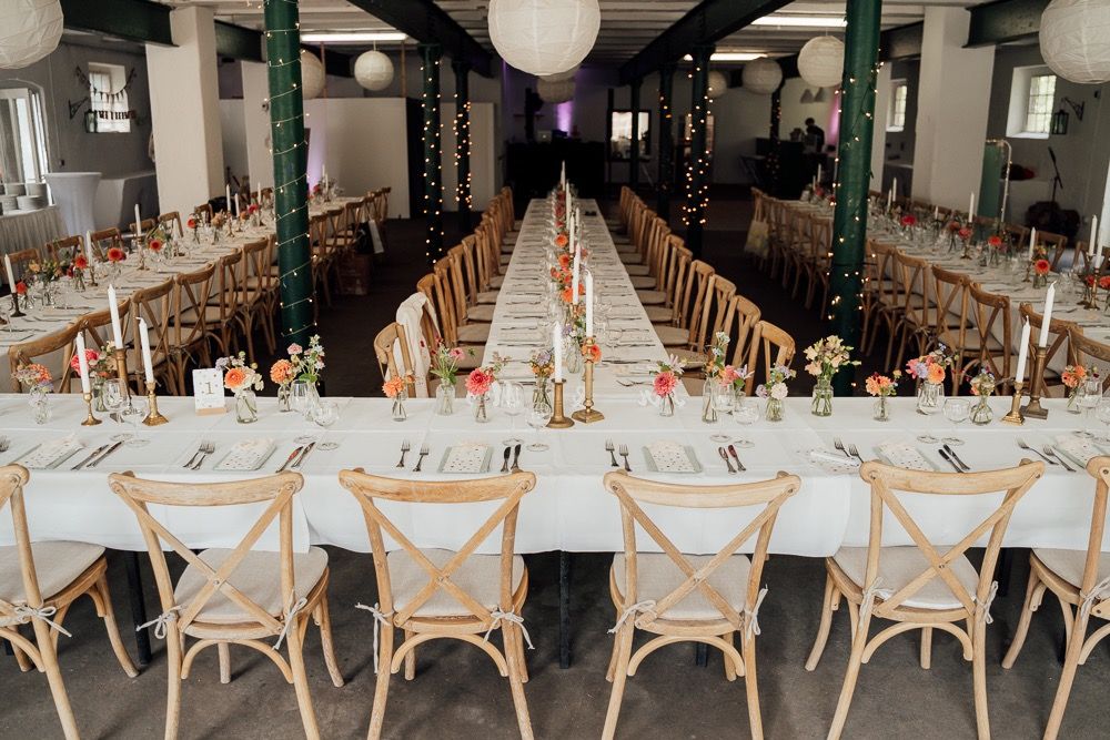 Long banquet tables with cross-back chairs set up for a wedding reception under hanging paper lanterns.