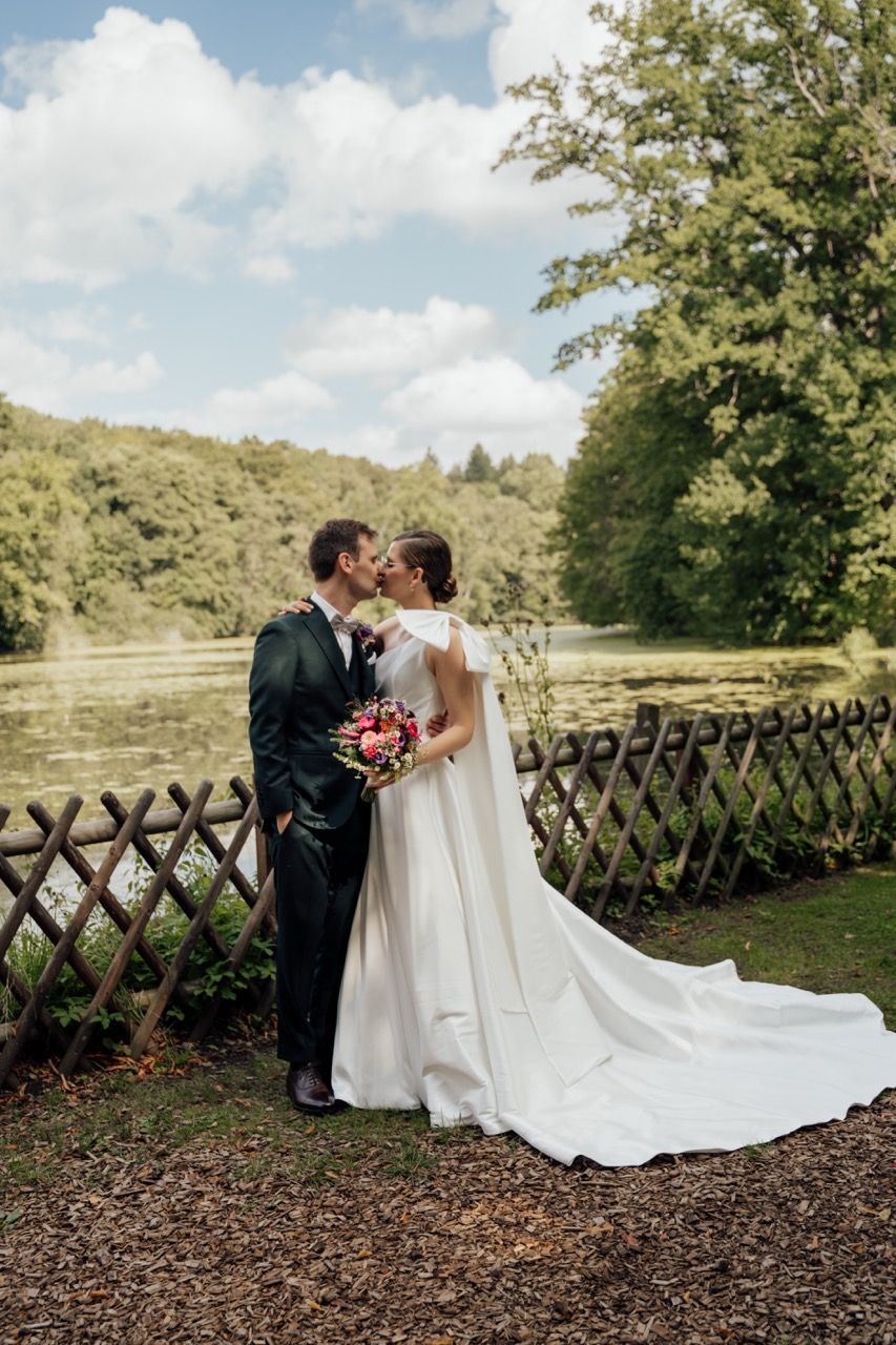 Bride and groom kissing in white wedding dress with cape train and dark suit by a lake with wooden fence.