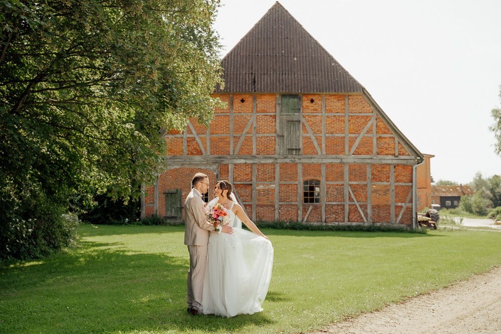 A couple in wedding attire stands on a lawn in front of a traditional German timber-framed barn building.