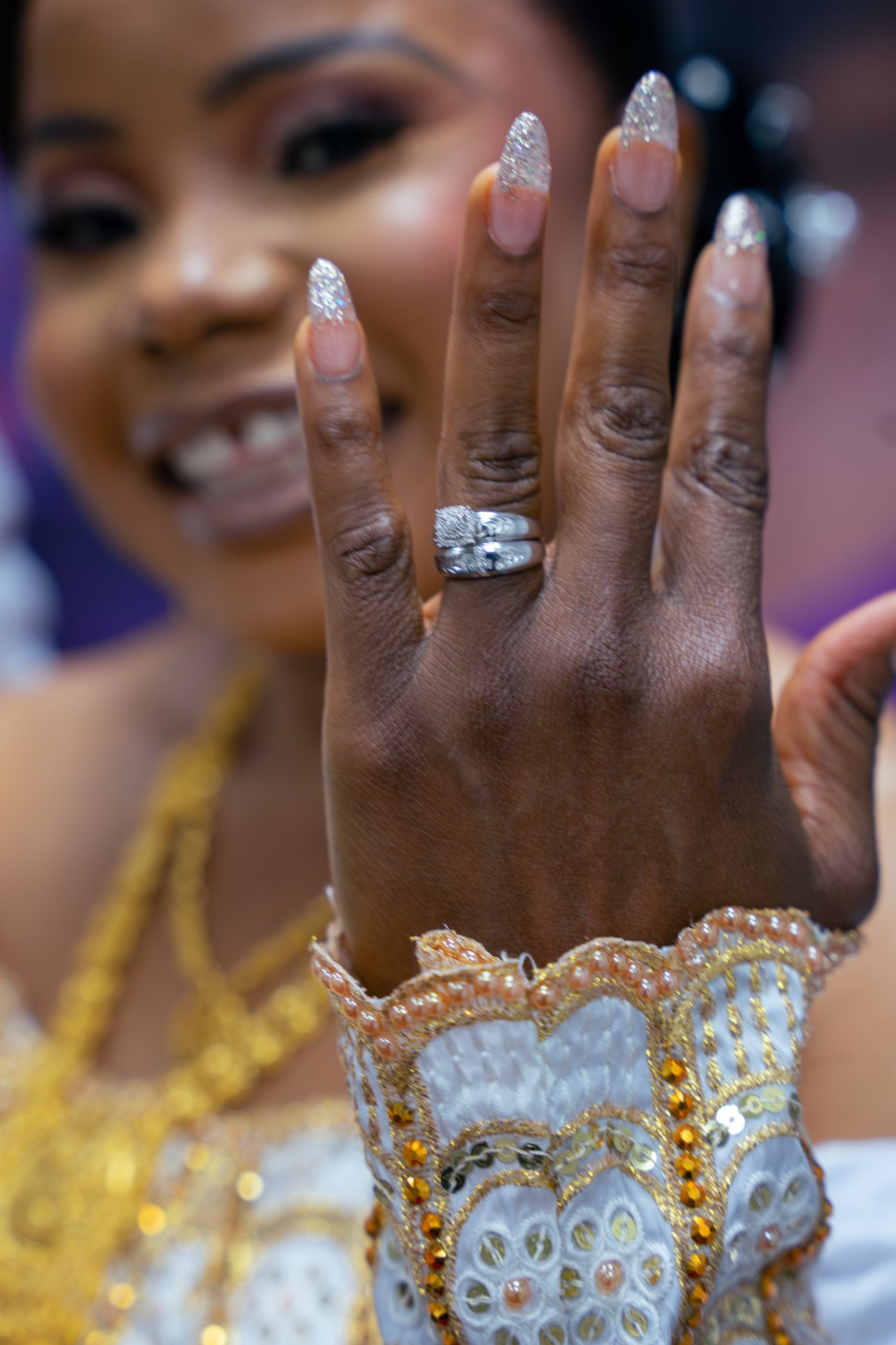 A close-up view of a sparkling diamond engagement ring on a hand against a white dress and gold necklace background.
