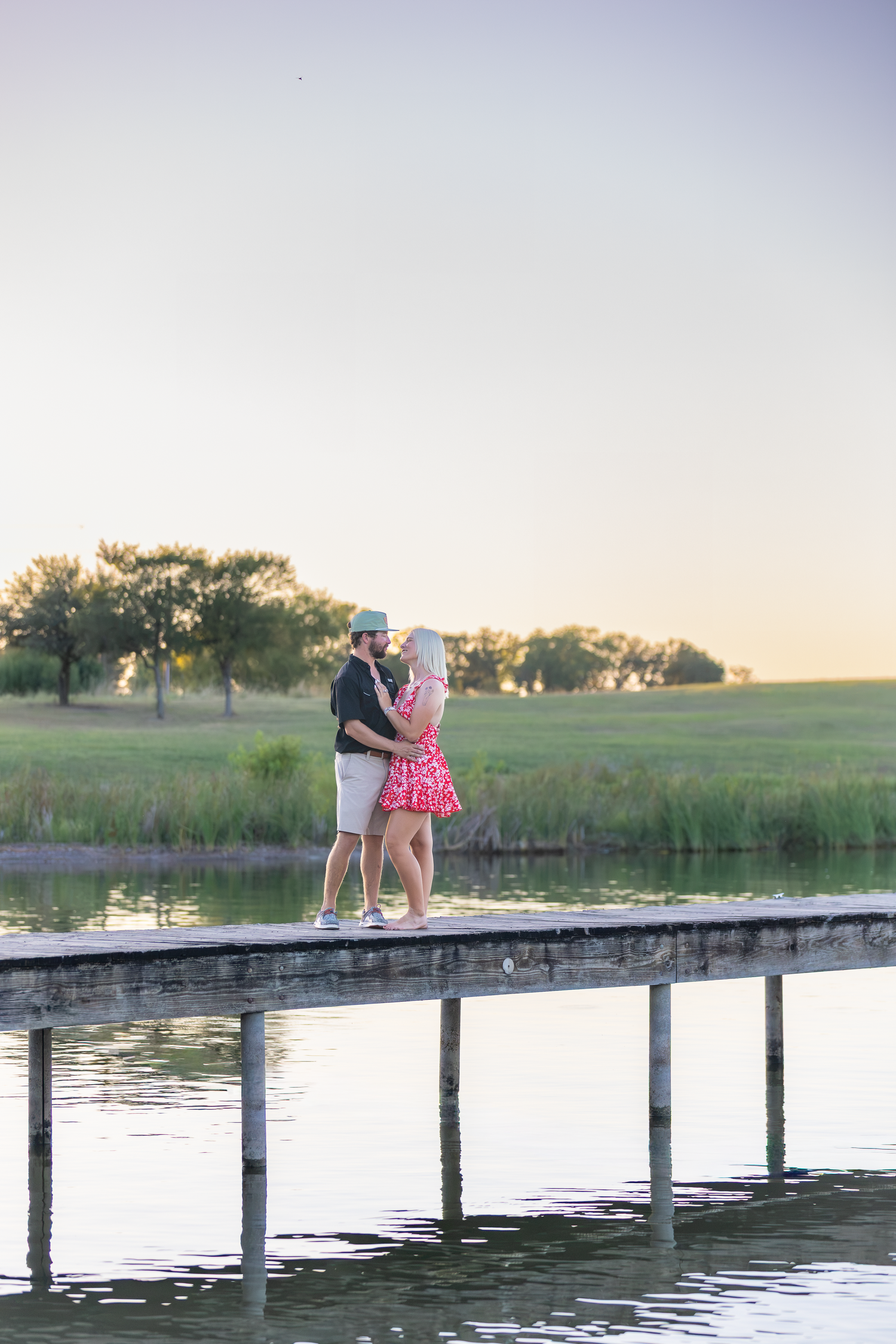 Couple embracing on a wooden dock by a serene lake at sunset, surrounded by lush green fields.