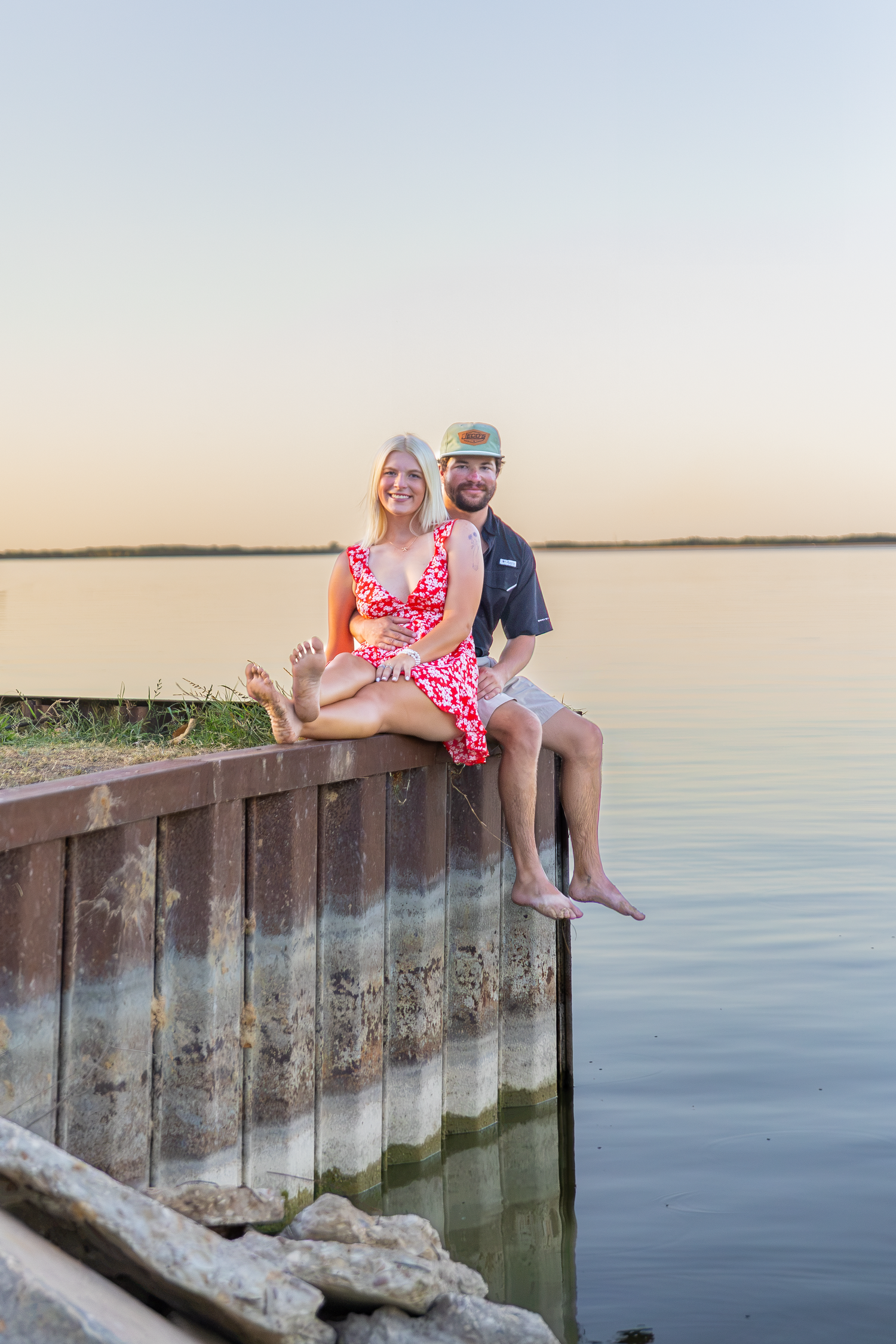Couple sitting together on a weathered dock wall by a calm lake at sunset, woman in red dress, man in cap.